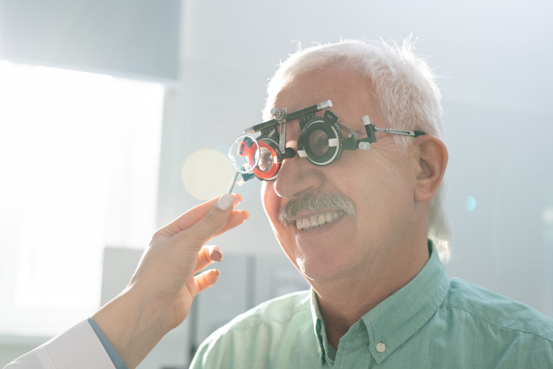 Smiling senior man sitting in front of ophthalmologist during medical check-up of his eyesight with optometrist trial frame in modern clinics